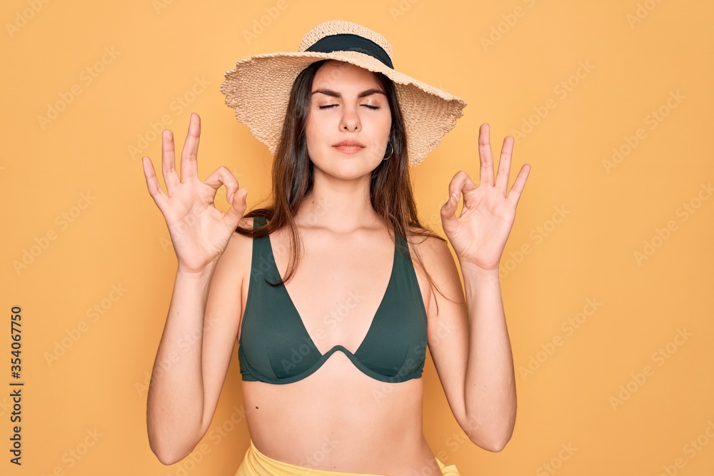 Young beautiful girl wearing swimwear bikini and summer sun hat over yellow background relax and smiling with eyes closed doing meditation gesture with fingers. Yoga concept. Young beautiful girl wearing swimwear bikini and summer sun hat over yellow background relax and smiling with eyes closed doing meditation gesture with fingers. Yoga concept.