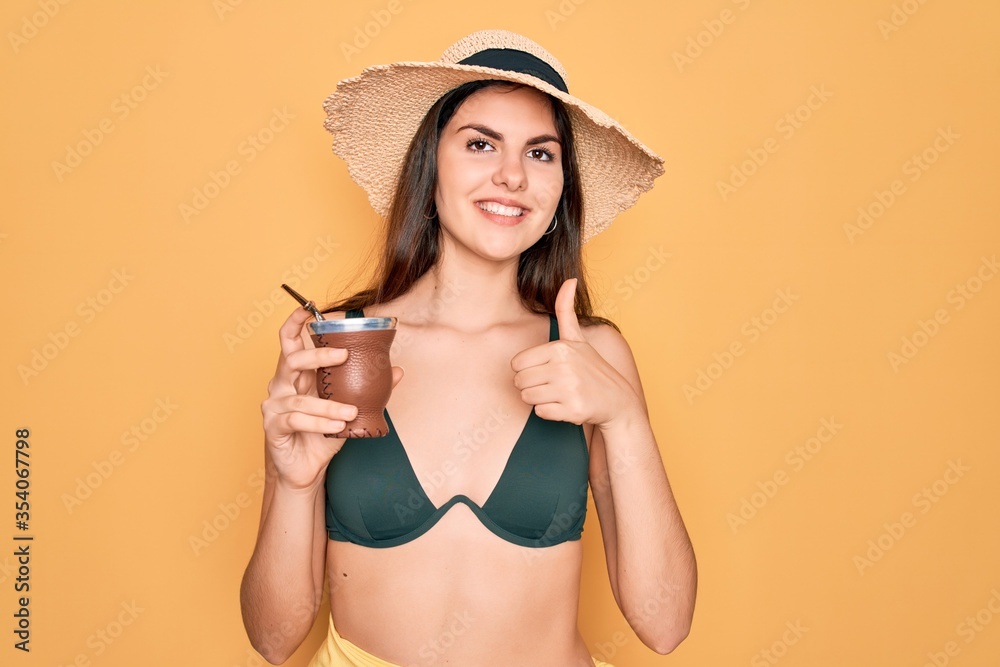 Young beautiful girl wearing swimwear bikini and summer hat drinking traditional mate drink happy with big smile doing ok sign, thumb up with fingers, excellent sign Young beautiful girl wearing swimwear bikini and summer hat drinking traditional mate drink happy with big smile doing ok sign, thumb up with fingers, excellent sign