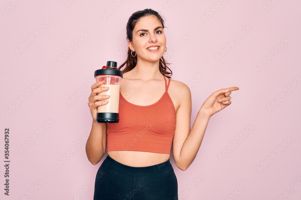 Young beautiful fitness woman wearing sport excersie clothes drinking protein shake very happy pointing with hand and finger to the side Young beautiful fitness woman wearing sport excersie clothes drinking protein shake very happy pointing with hand and finger to the side