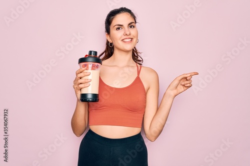 Young beautiful fitness woman wearing sport excersie clothes drinking protein shake very happy pointing with hand and finger to the side