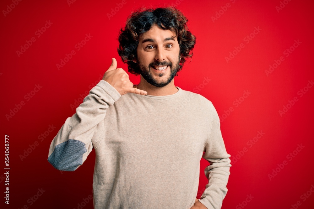 Young handsome man with beard wearing casual sweater standing over red background smiling doing phone gesture with hand and fingers like talking on the telephone. Communicating concepts.