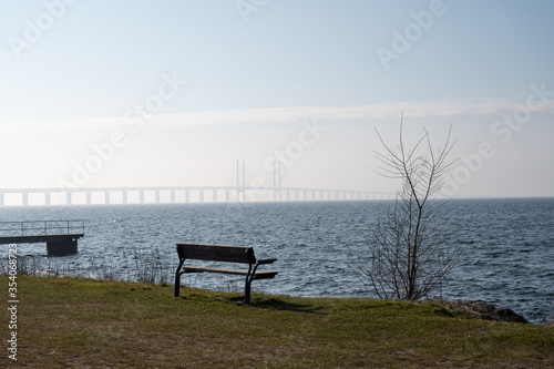 An empty bench just close to the sea with the Oresund bridge in the fog in the background