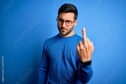 Young handsome man with beard wearing casual sweater and glasses over blue background Showing middle finger, impolite and rude fuck off expression © Krakenimages.com