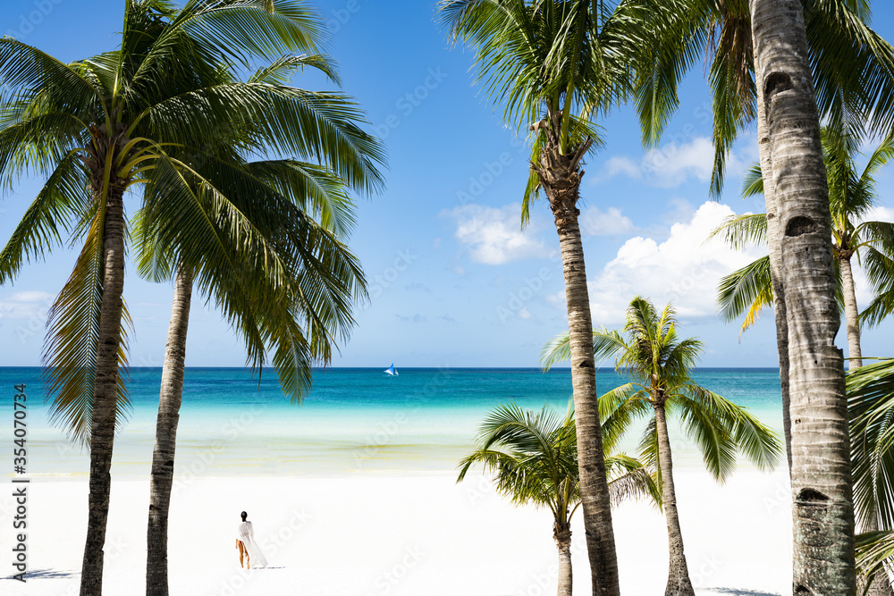 (Selective focus) Stunning view of woman walking on a white sand beach bathed by a turquoise sea, beautiful coconut palm trees in the foreground. White Beach, Boracay Island, Philippines.
