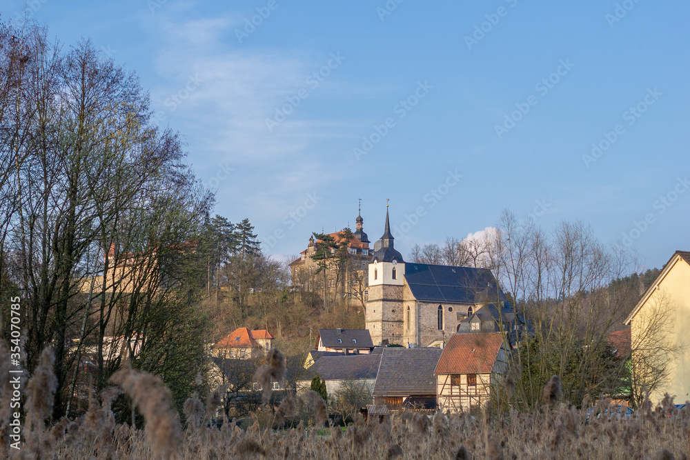 Dreifaltigkeitskirche mit Turm aus Schiefer am Schlossberg in Neuhaus-Schierschnitz in Thüringen. 
Kirche am Berg neben einer Burg bei blauem Himmel mit Bäumen und Steinhäusern.