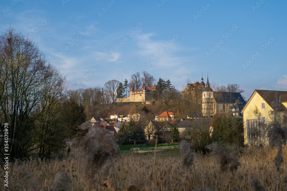 Burg Neuhaus am Schlossberg in Neuhaus-Schierschnitz in Thüringen neben der Dreifaltigkeitskirche. Landschaftsaufnahme einer kleinen Ortschaft mit Kirchturm und Burg am Berg bei Tag.