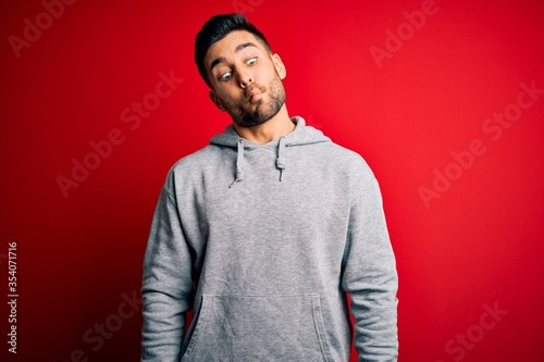 Young handsome sportsman wearing sweatshirt standing over isolated red background making fish face with lips, crazy and comical gesture. Funny expression.