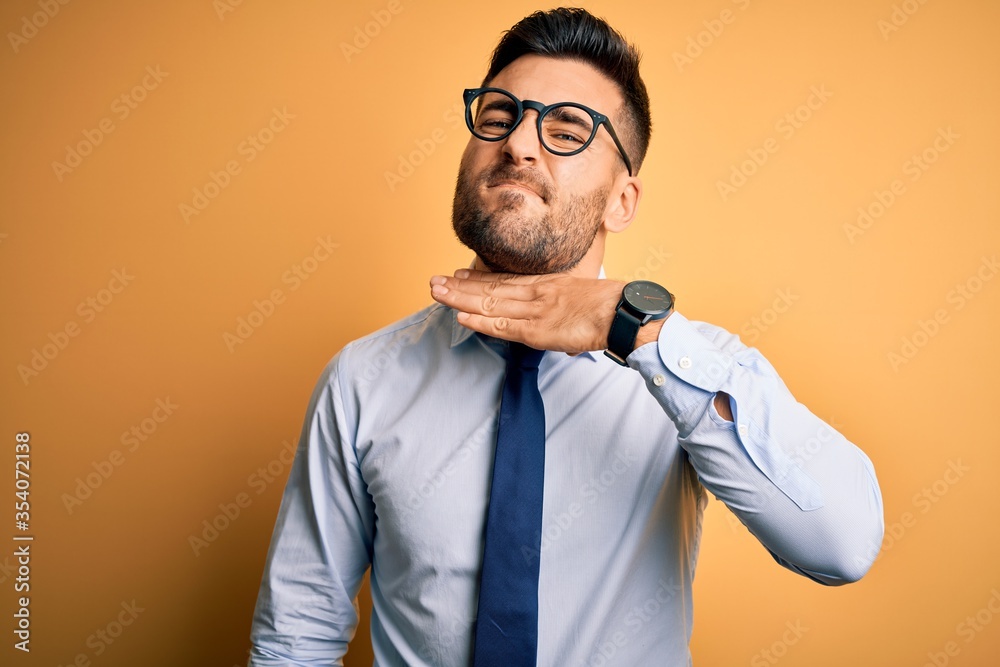 Young handsome businessman wearing tie and glasses standing over yellow background cutting throat with hand as knife, threaten aggression with furious violence Young handsome businessman wearing tie and glasses standing over yellow background cutting throat with hand as knife, threaten aggression with furious violence
