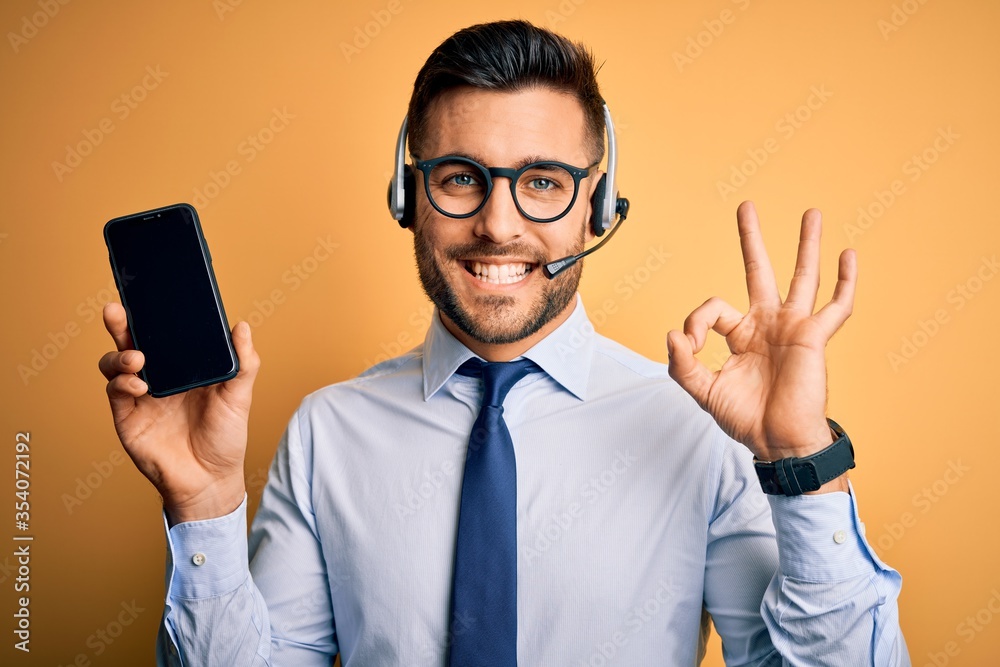 Business operator man with customer service headset from call center showing smartphone screen doing ok sign with fingers, excellent symbol Business operator man with customer service headset from call center showing smartphone screen doing ok sign with fingers, excellent symbol