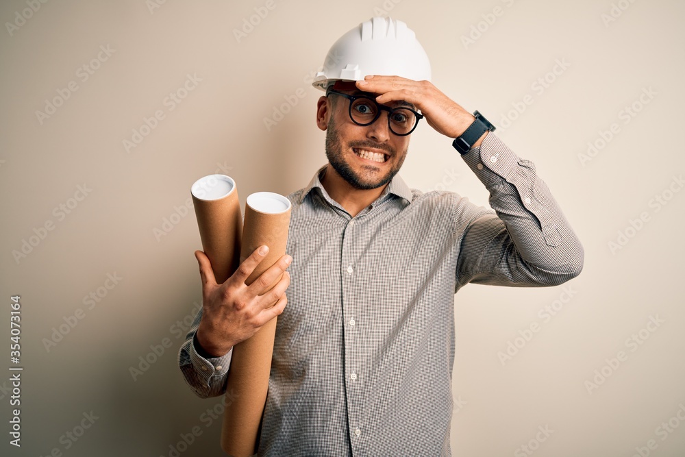 Young architect man wearing contractor helmet holding project paper plan over isolated background stressed with hand on head, shocked with shame and surprise face, angry and frustrated. Fear and upset Young architect man wearing contractor helmet holding project paper plan over isolated background stressed with hand on head, shocked with shame and surprise face, angry and frustrated. Fear and upset