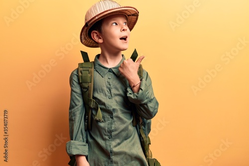 Cute blond kid wearing explorer hat and backpack pointing thumb up to the side smiling happy with open mouth photo