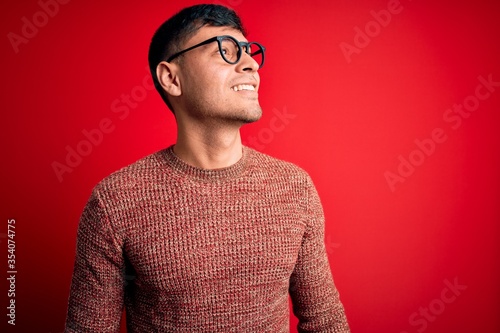 Young handsome hispanic man wearing nerd glasses over red background looking away to side with smile on face, natural expression. Laughing confident.