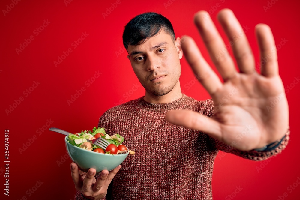 Young hispanic man eating vegetarian healthy salad over red isolated background with open hand doing stop sign with serious and confident expression, defense gesture Young hispanic man eating vegetarian healthy salad over red isolated background with open hand doing stop sign with serious and confident expression, defense gesture