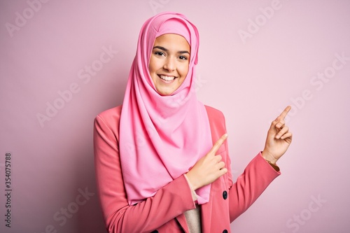 Young beautiful girl wearing muslim hijab standing over isolated pink background smiling and looking at the camera pointing with two hands and fingers to the side.