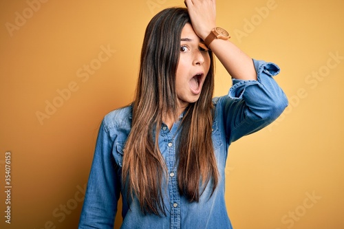 Young beautiful girl wearing casual denim shirt standing over isolated yellow background surprised with hand on head for mistake, remember error. Forgot, bad memory concept.