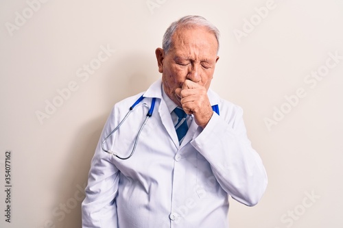 Senior grey-haired doctor man wearing coat and stethoscope standing over white background feeling unwell and coughing as symptom for cold or bronchitis. Health care concept.