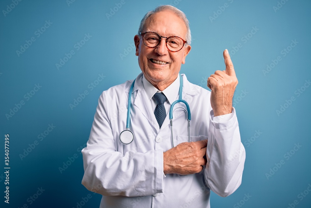 Senior grey haired doctor man wearing stethoscope and medical coat over blue background with a big smile on face, pointing with hand and finger to the side looking at the camera. Senior grey haired doctor man wearing stethoscope and medical coat over blue background with a big smile on face, pointing with hand and finger to the side looking at the camera.