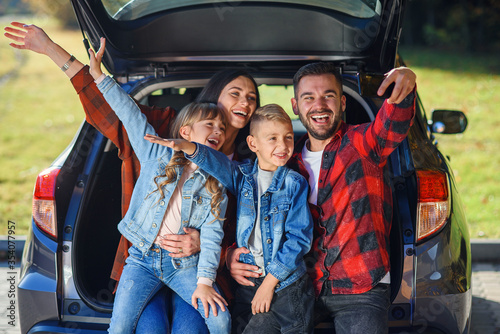 Happy stylish parents with their cute lovely children are making funny selfie on smart phone while sitting in the trunk. Happy modern family concept.