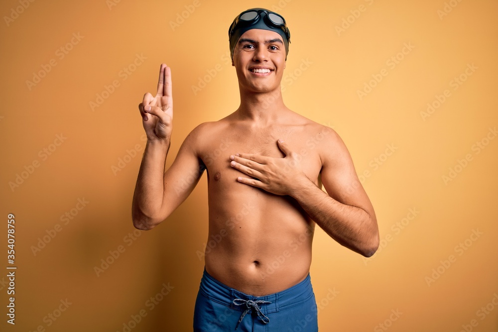 Young handsome man shirtless wearing swimsuit and swim cap over isolated yellow background smiling swearing with hand on chest and fingers up, making a loyalty promise oath