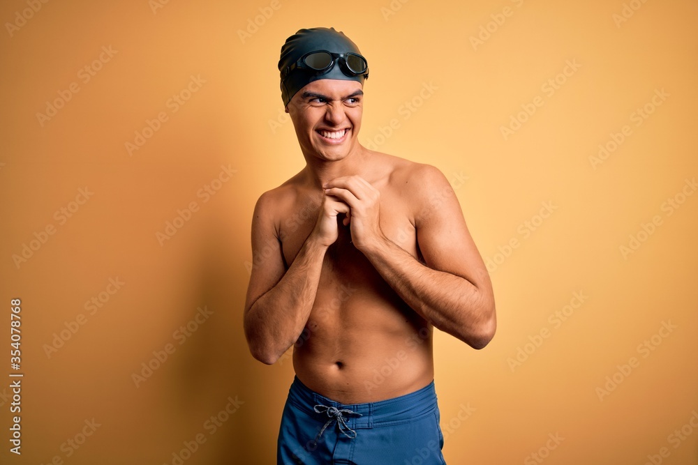 Young handsome man shirtless wearing swimsuit and swim cap over isolated yellow background laughing nervous and excited with hands on chin looking to the side