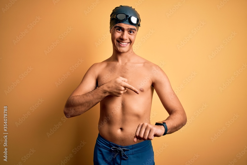 Young handsome man shirtless wearing swimsuit and swim cap over isolated yellow background In hurry pointing to watch time, impatience, upset and angry for deadline delay