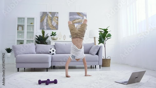 Good-looking sportive confident teen boy doing handstand on the carpet,walking on his hands and falling down on the couch
