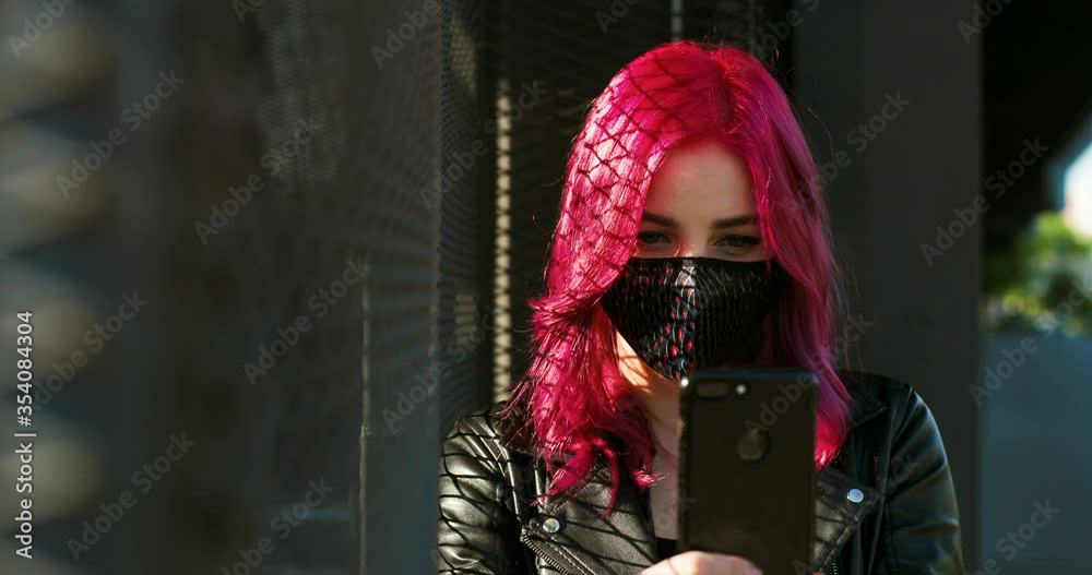 Caucasian young stylish hipster with pink hair tapping or texting message on smartphone outdoors. Extraordinary girl in mask scrolling on mobile phone. Female using cellphone. Covid-19 pandemic.