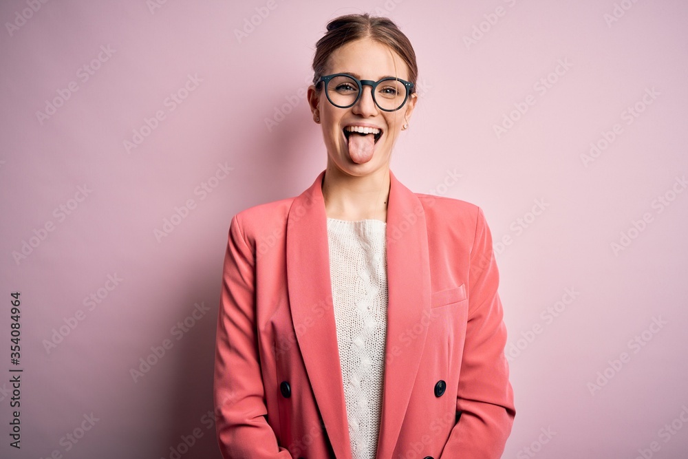 Young beautiful redhead woman wearing jacket and glasses over isolated pink background sticking tongue out happy with funny expression. Emotion concept.