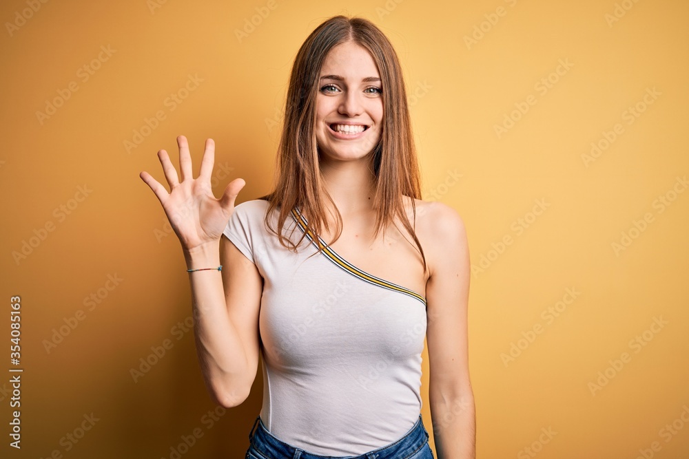 Young beautiful redhead woman wearing casual t-shirt over isolated yellow background showing and pointing up with fingers number five while smiling confident and happy. Young beautiful redhead woman wearing casual t-shirt over isolated yellow background showing and pointing up with fingers number five while smiling confident and happy.
