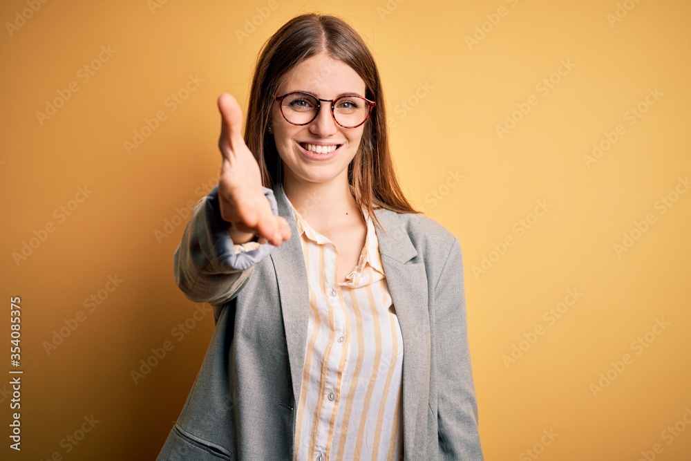 Young beautiful redhead woman wearing jacket and glasses over isolated yellow background smiling friendly offering handshake as greeting and welcoming. Successful business.
