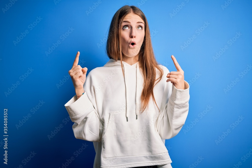 Young beautiful redhead sporty woman wearing sweatshirt over isolated blue background amazed and surprised looking up and pointing with fingers and raised arms. Young beautiful redhead sporty woman wearing sweatshirt over isolated blue background amazed and surprised looking up and pointing with fingers and raised arms.