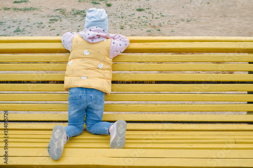 lonely child on a background of a yellow park bench. girl in a bright jacket, white hat and blue jeans. insulation. concept of loneliness, self-isolation © moisevich