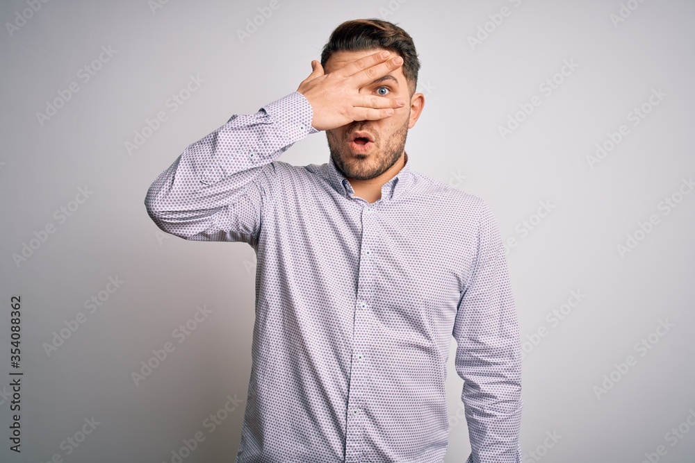 Young business man with blue eyes standing over isolated background peeking in shock covering face and eyes with hand, looking through fingers with embarrassed expression. Young business man with blue eyes standing over isolated background peeking in shock covering face and eyes with hand, looking through fingers with embarrassed expression.