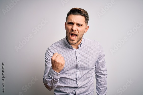 Young business man with blue eyes standing over isolated background angry and mad raising fist frustrated and furious while shouting with anger. Rage and aggressive concept.