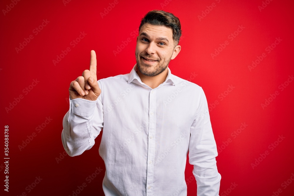 Fototapeta premium Young business man with blue eyes wearing elegant shirt standing over red isolated background showing and pointing up with finger number one while smiling confident and happy.