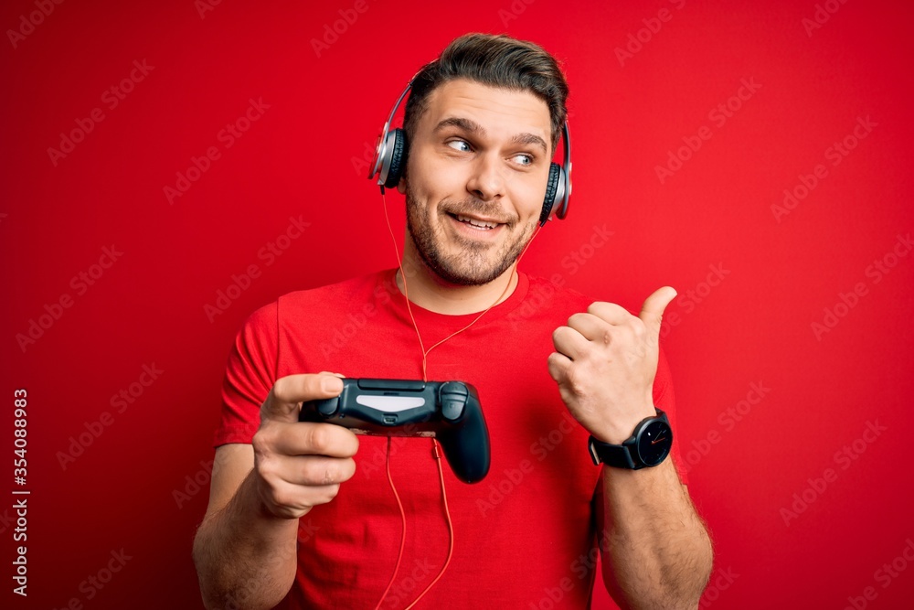 Young gamer man with blue eyes playing video games using gamepad joystick pointing and showing with thumb up to the side with happy face smiling Young gamer man with blue eyes playing video games using gamepad joystick pointing and showing with thumb up to the side with happy face smiling