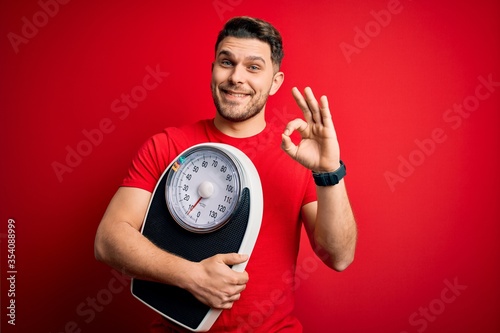 Young fitness man with blue eyes holding scale dieting for healthy weight over red background doing ok sign with fingers, excellent symbol