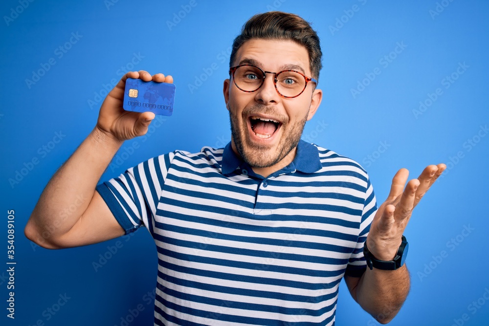 Young man with blue eyes wearing glasses and holding credit card over blue background very happy and excited, winner expression celebrating victory screaming with big smile and raised hands Young man with blue eyes wearing glasses and holding credit card over blue background very happy and excited, winner expression celebrating victory screaming with big smile and raised hands