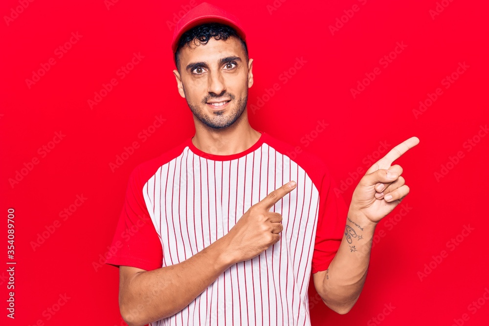 Young hispanic man wearing baseball uniform smiling and looking at the camera pointing with two hands and fingers to the side. Young hispanic man wearing baseball uniform smiling and looking at the camera pointing with two hands and fingers to the side.