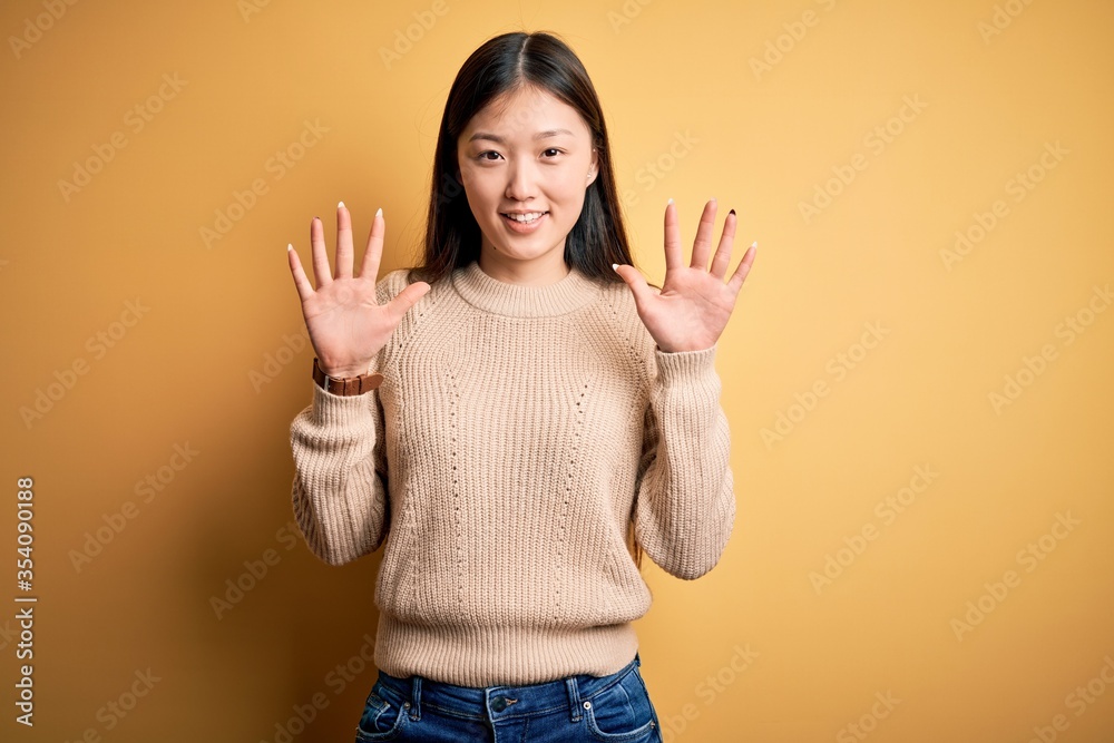 Young beautiful asian woman wearing casual sweater over yellow isolated background showing and pointing up with fingers number ten while smiling confident and happy.