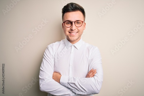 Young handsome business mas wearing glasses and elegant shirt over isolated background happy face smiling with crossed arms looking at the camera. Positive person.