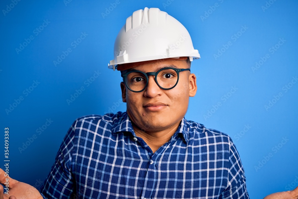 Young handsome engineer latin man wearing safety helmet over isolated blue background clueless and confused expression with arms and hands raised. Doubt concept.