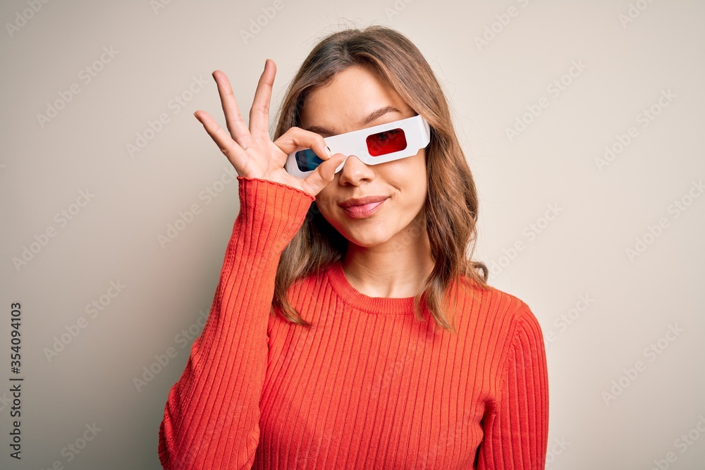 Young blonde girl wearing 3d cinema glasses over isolated background doing ok gesture with hand smiling, eye looking through fingers with happy face. Young blonde girl wearing 3d cinema glasses over isolated background doing ok gesture with hand smiling, eye looking through fingers with happy face.