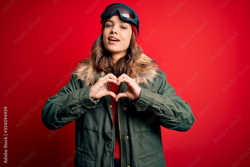 Young blonde girl wearing ski glasses and winter coat for ski weather over red background smiling in love doing heart symbol shape with hands. Romantic concept. Young blonde girl wearing ski glasses and winter coat for ski weather over red background smiling in love doing heart symbol shape with hands. Romantic concept.