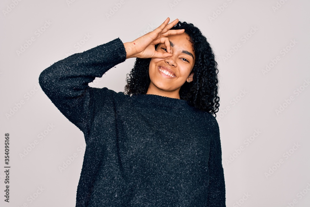 Young african american curly woman wearing casual sweater standing over white background doing ok gesture with hand smiling, eye looking through fingers with happy face. Young african american curly woman wearing casual sweater standing over white background doing ok gesture with hand smiling, eye looking through fingers with happy face.