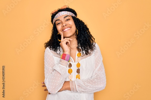 Young african american curly hippie woman wearing sunglasses and vintage accessories looking confident at the camera with smile with crossed arms and hand raised on chin. Thinking positive.