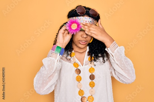 Young african american curly hippie woman wearing sunglasses holding pink flower on eye stressed with hand on head, shocked with shame and surprise face, angry and frustrated. Fear and upset