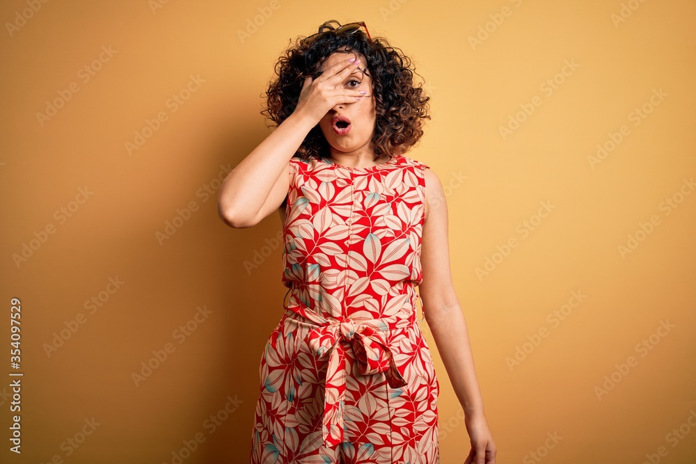 Young beautiful curly arab woman on vacation wearing summer floral dress and sunglasses peeking in shock covering face and eyes with hand, looking through fingers with embarrassed expression. Young beautiful curly arab woman on vacation wearing summer floral dress and sunglasses peeking in shock covering face and eyes with hand, looking through fingers with embarrassed expression.