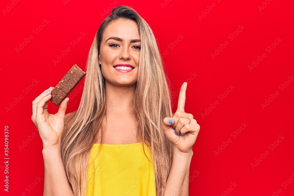Young beautiful blonde woman eating chocolate protein bar over isolated red background smiling with an idea or question pointing finger with happy face, number one Young beautiful blonde woman eating chocolate protein bar over isolated red background smiling with an idea or question pointing finger with happy face, number one