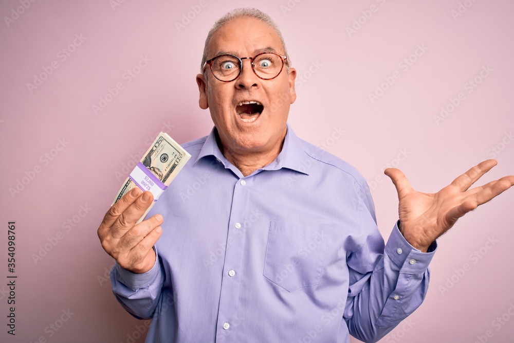 Middle age hoary man holding bunch of dollars banknotes over isolated pink background very happy and excited, winner expression celebrating victory screaming with big smile and raised hands Middle age hoary man holding bunch of dollars banknotes over isolated pink background very happy and excited, winner expression celebrating victory screaming with big smile and raised hands